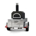 A portable outdoor pizza oven with a solar black finish, mounted on a trailer with a diamond plate pattern. It features a visible propane burner and a closed door at the front.